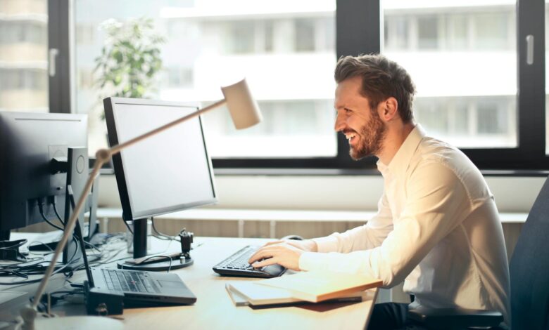 man in white dress shirt sitting on black rolling chair while facing black computer set and smiling